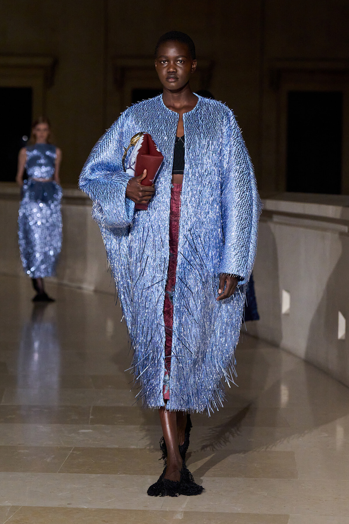 Model on a runway wearing a light-blue fringed coat, black top, burgundy pants, and black fringed shoes, clutching a red purse and a patterned bag.