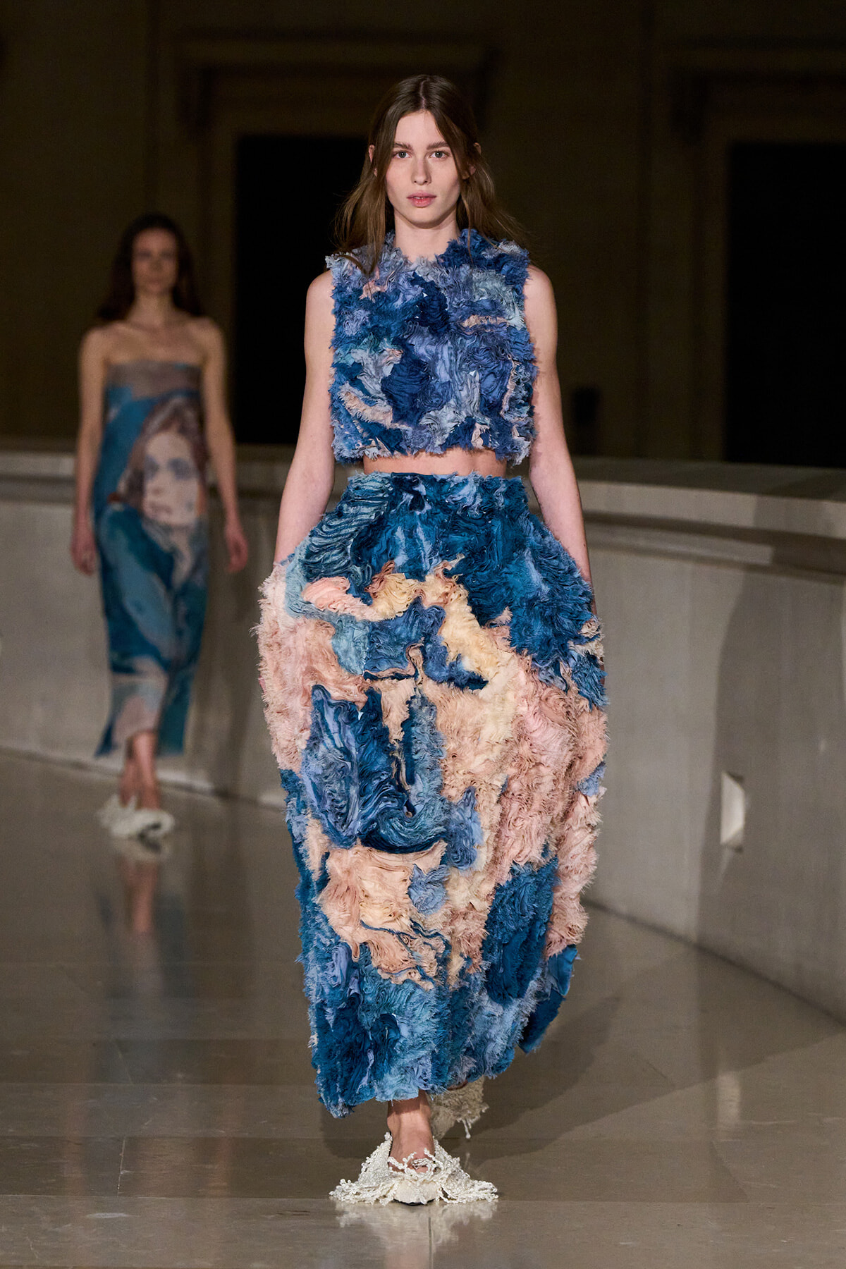 Model walking a runway in a two-piece textured fringe dress in blue and peach tones, with matching frayed footwear