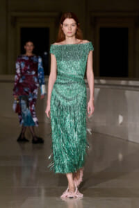 Model wearing an emerald green off-the-shoulder fringe gown walking a runway with a neutral expression.