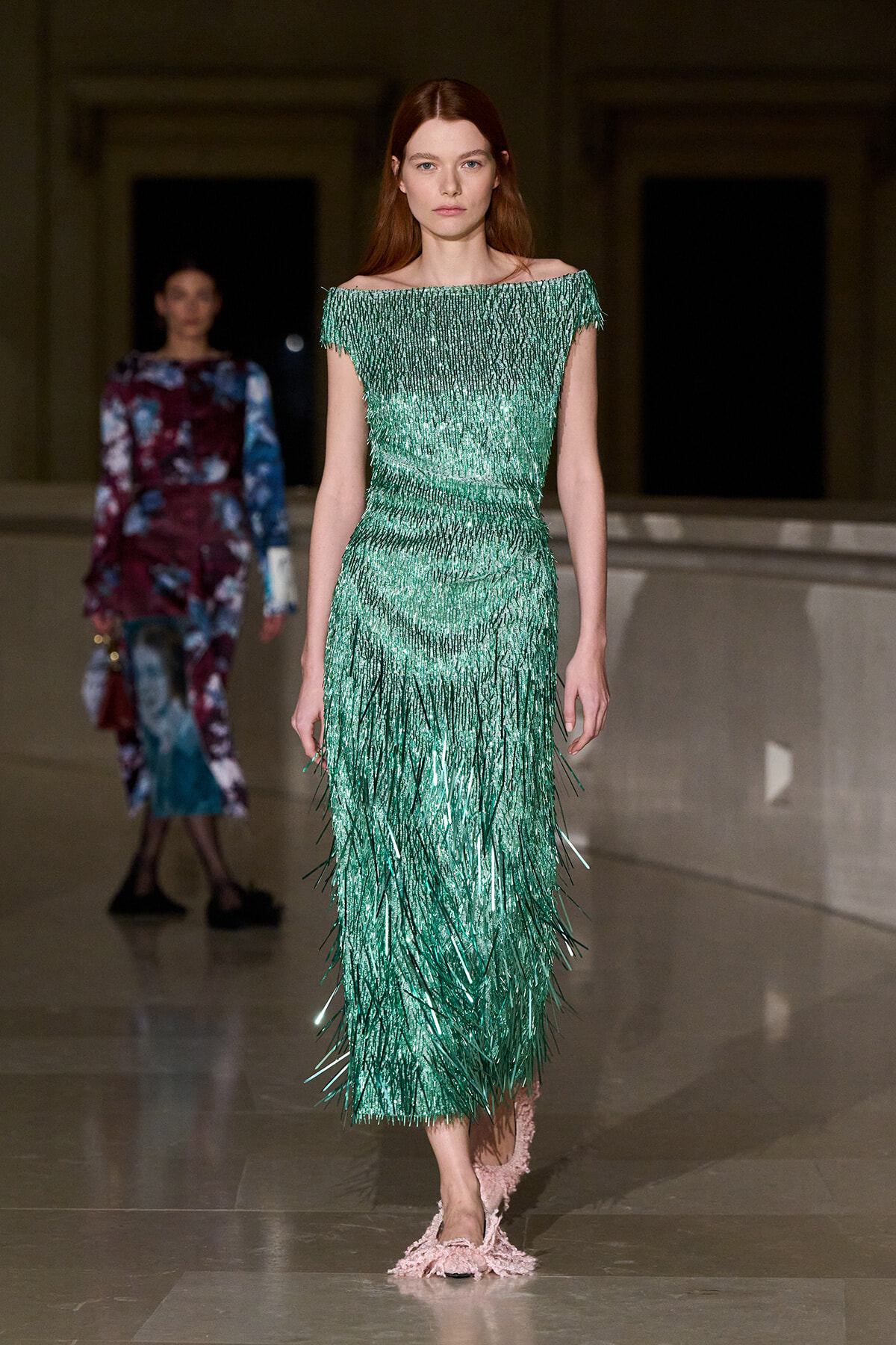 Model wearing an emerald green off-the-shoulder fringe gown walking a runway with a neutral expression.