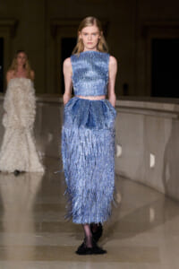 Model on a runway wearing a metallic blue fringe two-piece dress (crop top and long fringed skirt).