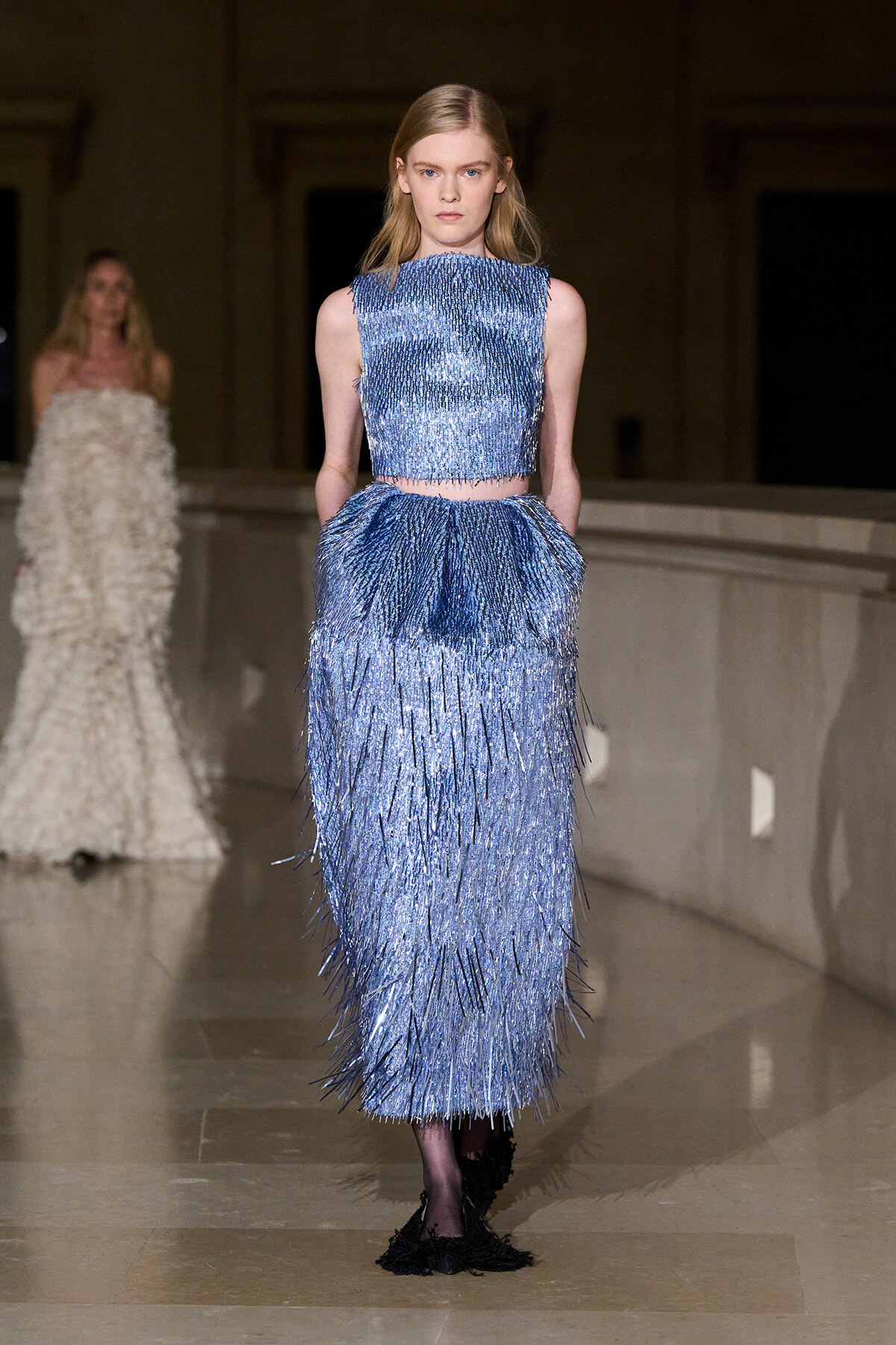 Model on a runway wearing a metallic blue fringe two-piece dress (crop top and long fringed skirt).