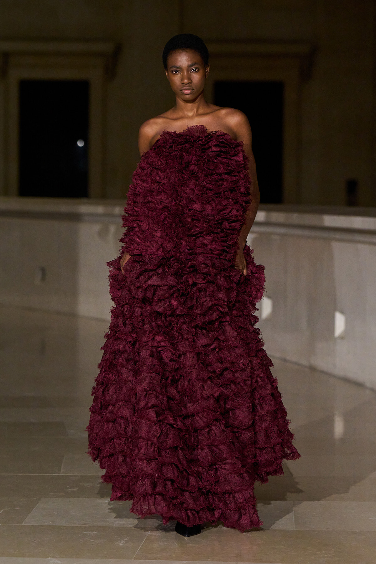 Model wearing a strapless burgundy textured gown with voluminous ruffles, posing indoors on a marble floor.