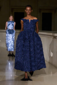 Runway model in a navy off-the-shoulder textured ballgown with a voluminous skirt; a sequined blue dress model stands in the background.