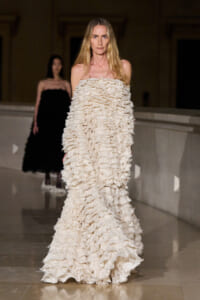 Model walking a runway in a floor-length off-shoulder cream ruffled gown; a second model in black is in the background.