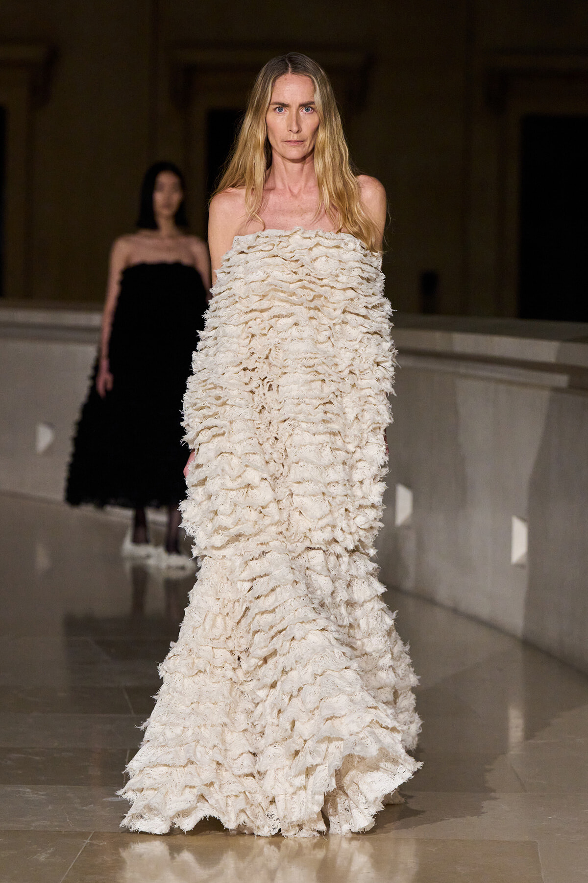 Model walking a runway in a floor-length off-shoulder cream ruffled gown; a second model in black is in the background.