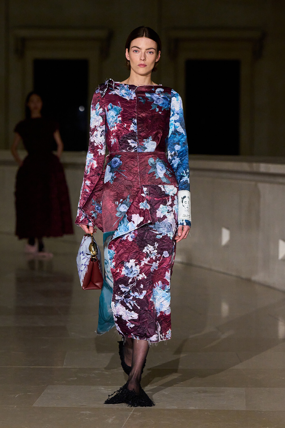 Model on the runway wears a long, crinkled floral-print dress in burgundy and blue tones, carrying a small red handbag.