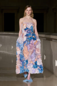 Model walking in a strapless, floor-length gown with voluminous, fluffy pastel petals in blue, pink, peach, and purple on a runway centering a modern venue.