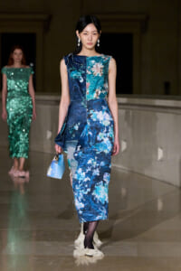Model walking a runway in a sleeveless, blue floral patchwork midi dress, carrying a small light-blue handbag.