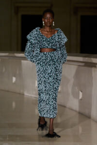Model on a runway wearing a teal textured two-piece outfit (cropped top and high-waisted skirt) with a fluffy fringe cardigan and spiky black heels, accessorized with drop earrings.
