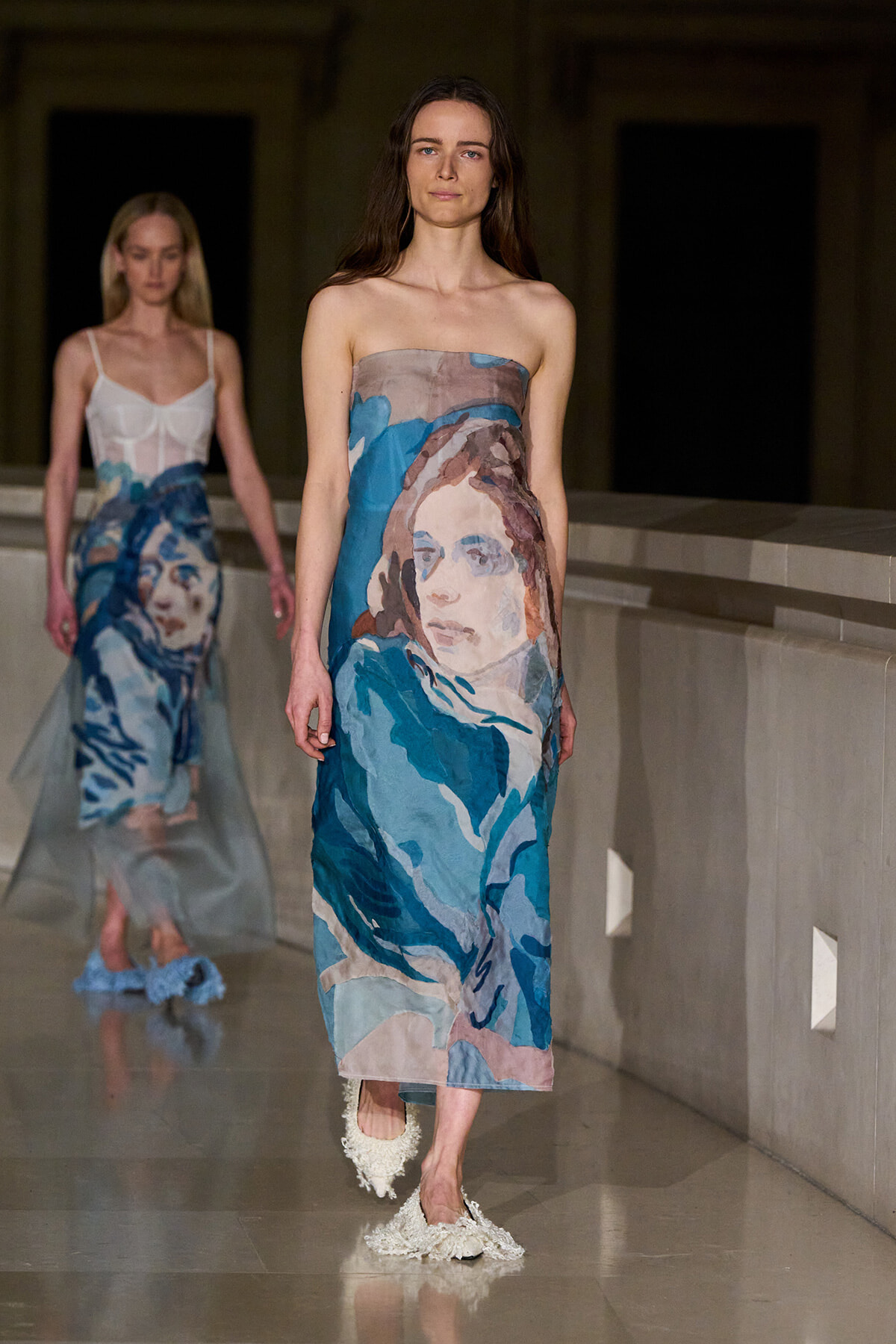Model walks the runway in a strapless dress with a large blue-toned portrait print on the fabric.