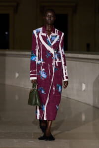 Model walking on runway in a burgundy floral long coat with white lapels, blue flowers, and a green textured handbag