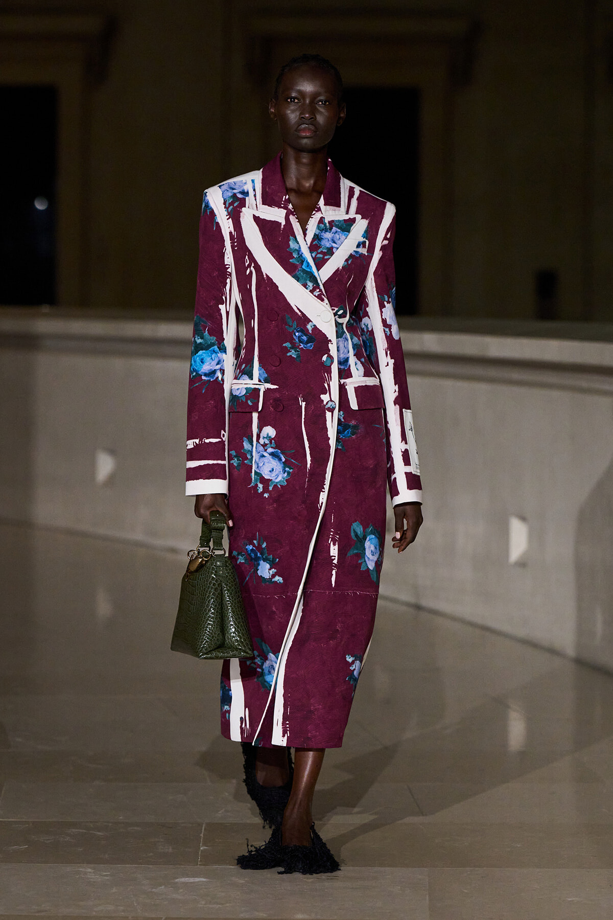 Model walking on runway in a burgundy floral long coat with white lapels, blue flowers, and a green textured handbag