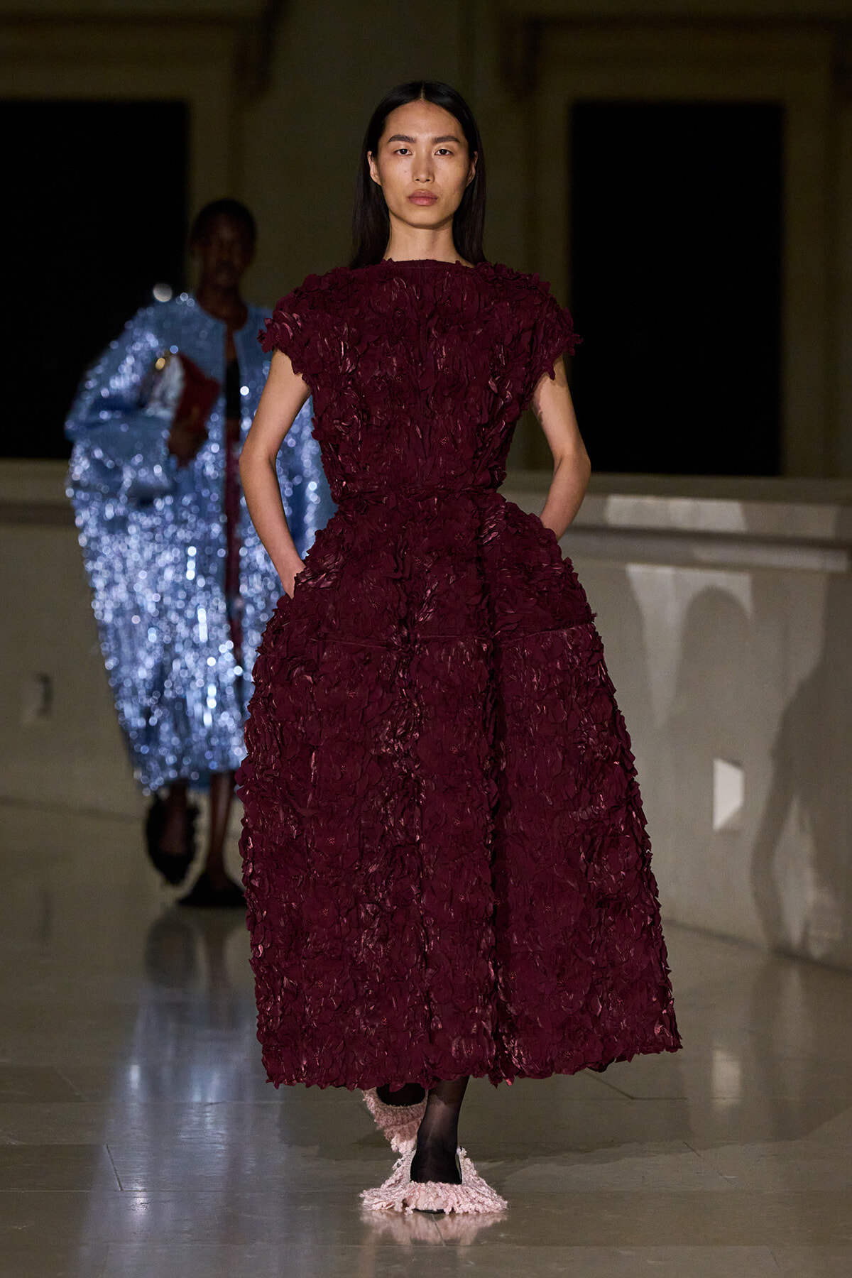 Model walks the runway in a voluminous burgundy textured dress, hands in pockets, neutral expression, dark hair framing her face.