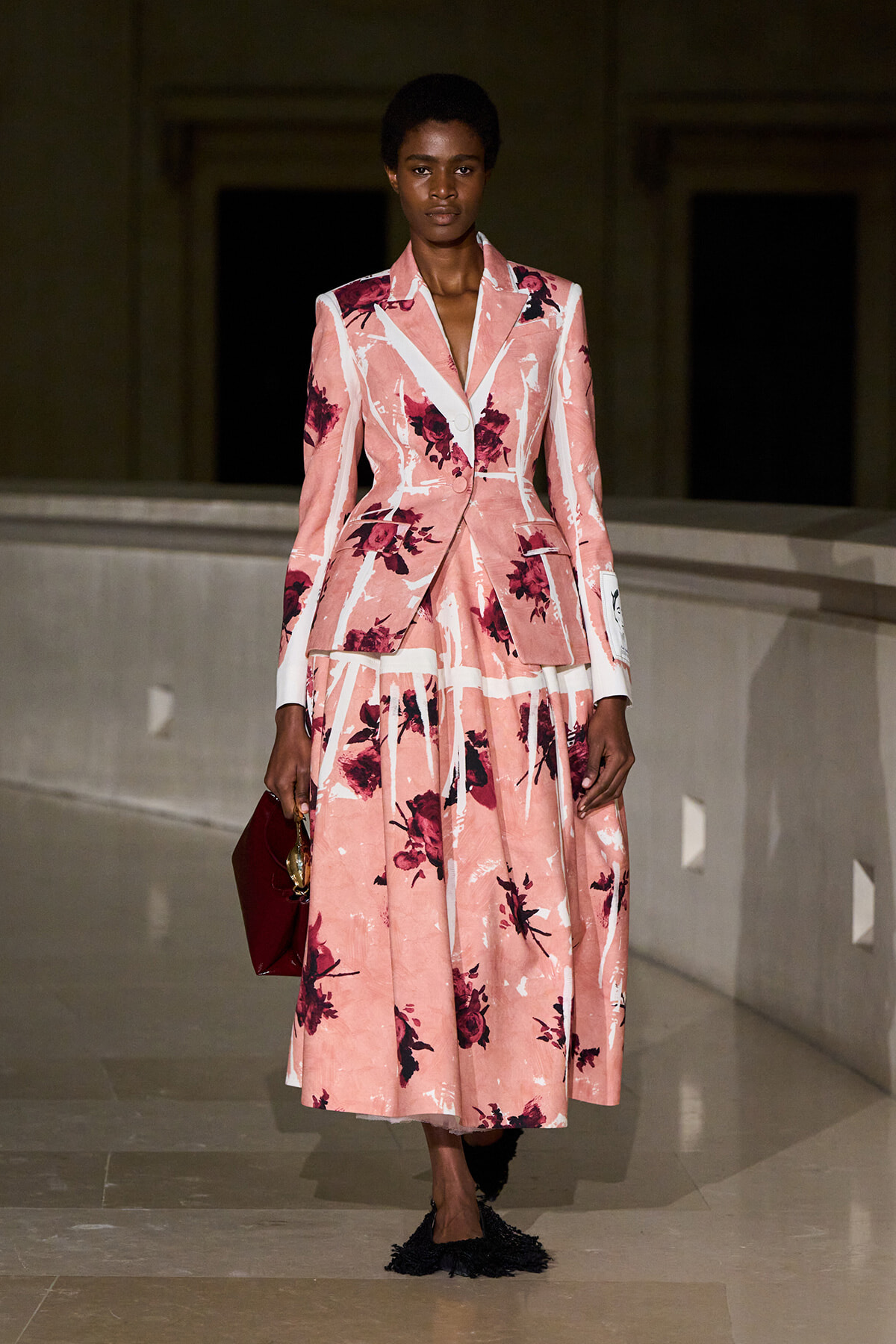 Model in a pink floral tailored blazer with matching A-line skirt, holding a burgundy clutch on a runway.