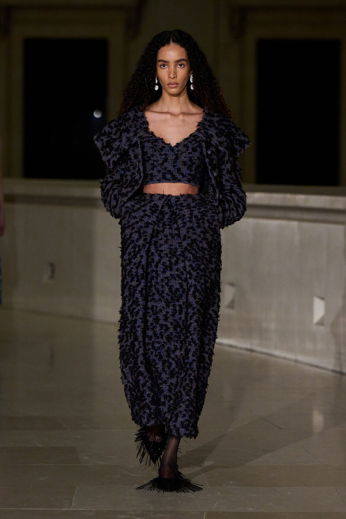 Model wearing a dark textured two-piece outfit with a cropped top and long skirt on a runway, dramatic oversized sleeves and statement earrings.