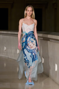 Model walking a runway in a sheer white-bodice gown with a large blue watercolor portrait skirt and light blue textured shoes