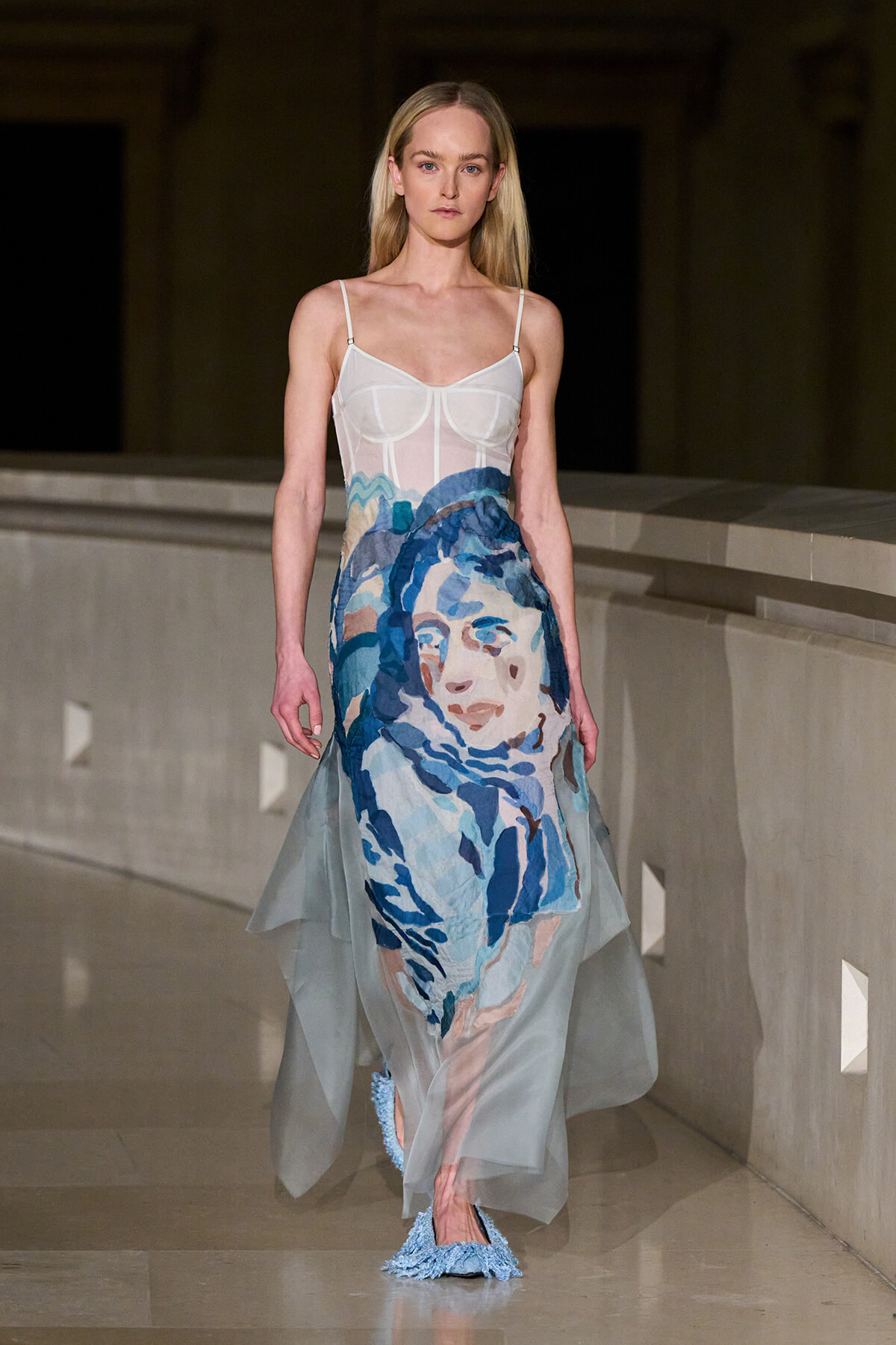 Model walking a runway in a sheer white-bodice gown with a large blue watercolor portrait skirt and light blue textured shoes