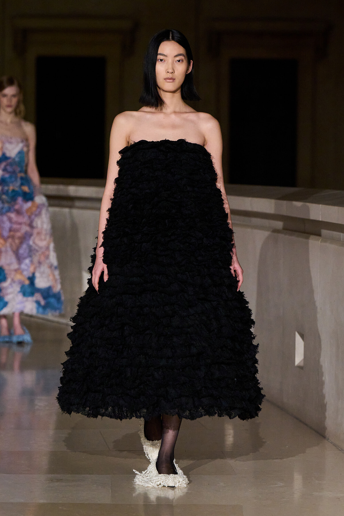 Model walking the runway in a strapless black textured gown with a voluminous skirt, dark hair, and a neutral expression.