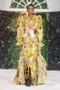 Model on a snowy runway wearing a yellow floral gown with puff sleeves, ruffles, and a pink rose belt.