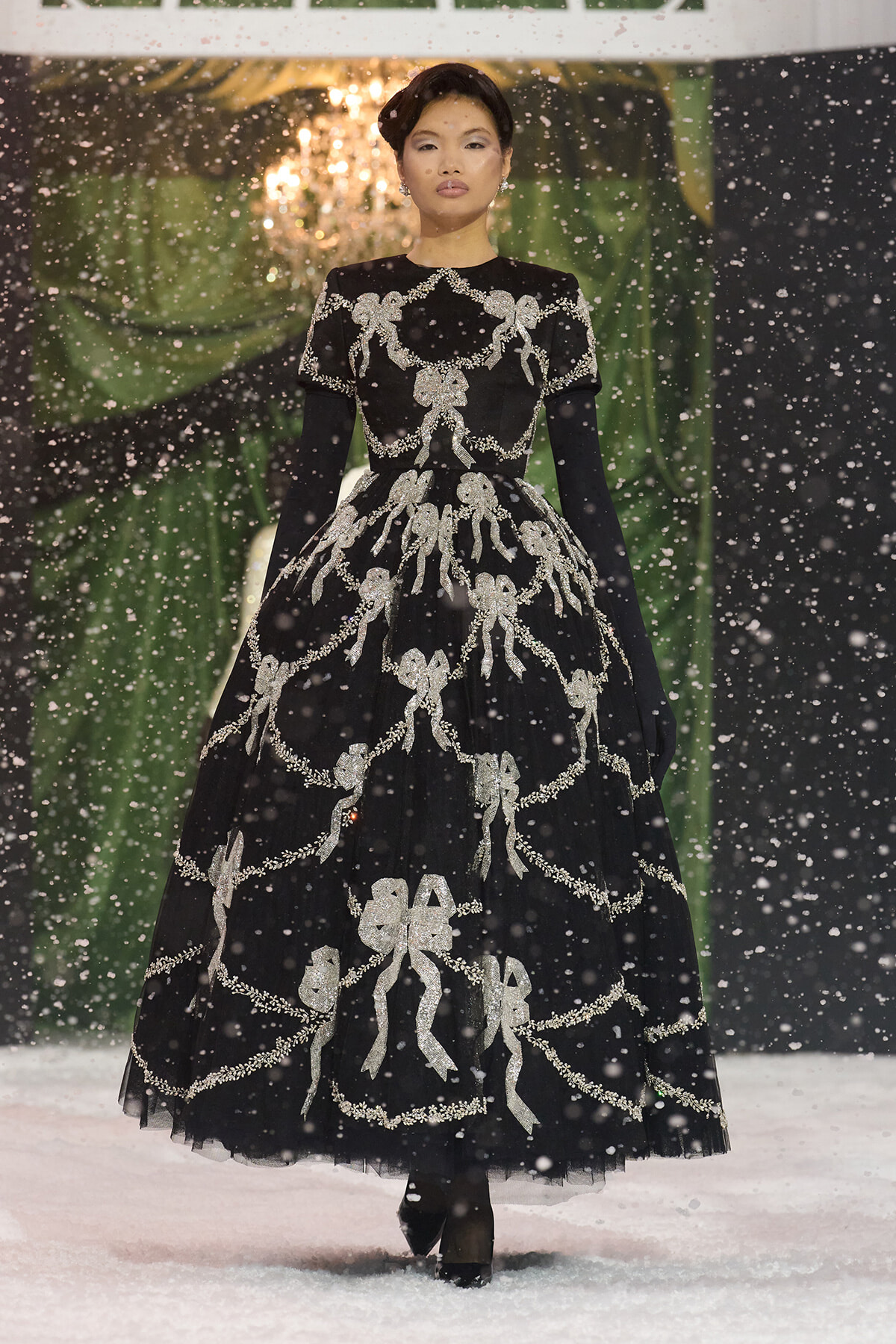 Model walking in a black floor-length gown adorned with silver bow and garland embroidery; snowfall on a fashion runway backdrop.
