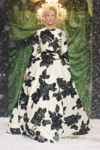 Model wearing a white ball gown with large black floral embroidery walking a snowy runway with a chandelier behind.