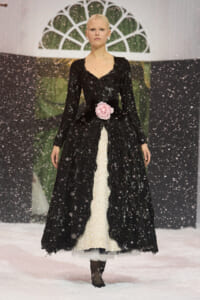 Model walking a snowy runway in a black lace gown with a pink rose belt, amid falling snowflakes.