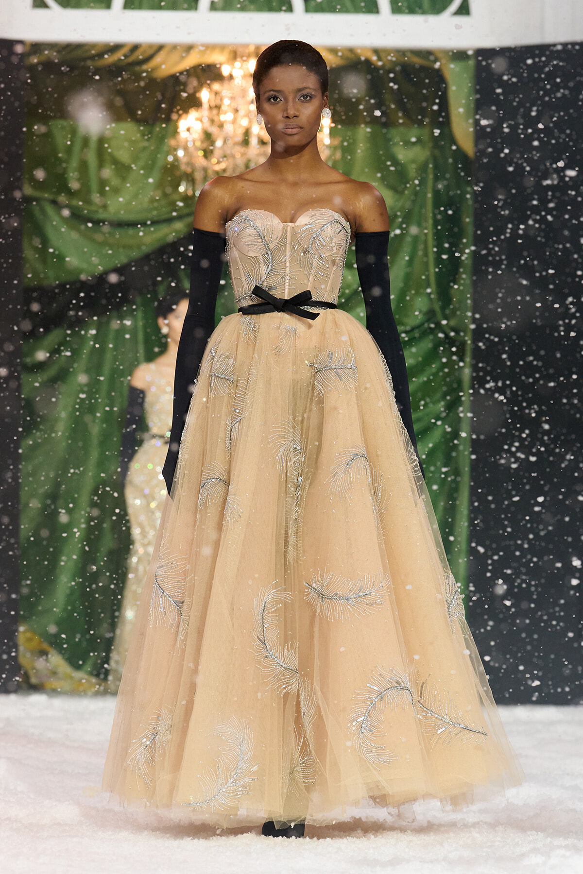 Model in a strapless champagne ballgown with silver embroidery, black opera gloves, and a black bow belt on a snowy runway backdrop.