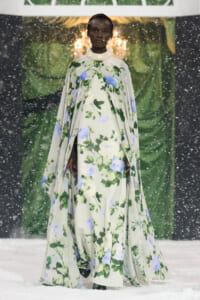 Model walks a flowing floral cape gown on a snowy runway, green and blue flower pattern against a pale background.