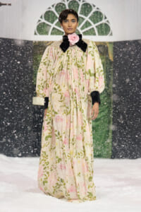 Model walking on a snowy runway in a long cream floral dress with pink rose prints, black velvet bow at the collar, pearl embellishments, and a pink rose at the neck.