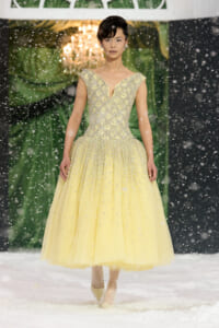Model walking a snowy runway in a pale yellow, bead-embellished ball gown with an off-the-shoulder V-neck and full tulle skirt, pale heels.
