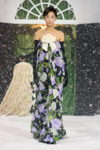 Model on a snowy runway wearing an off-shoulder floral gown with a large white bow and long black gloves, posing as she walks.