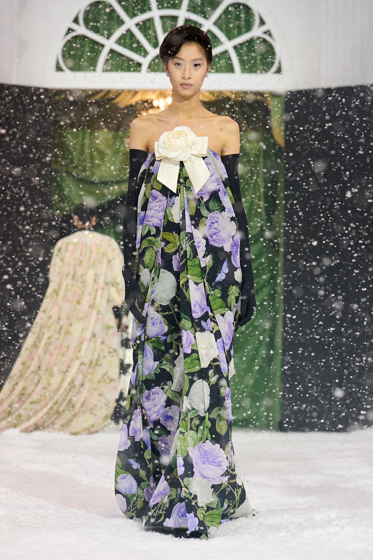 Model on a snowy runway wearing an off-shoulder floral gown with a large white bow and long black gloves, posing as she walks.