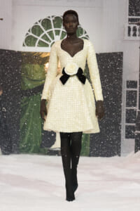 Model walking the runway in a cream beaded dress with a deep V neckline and a large white rose belt with black ribbons, snow-like backdrop and falling faux snow behind her.