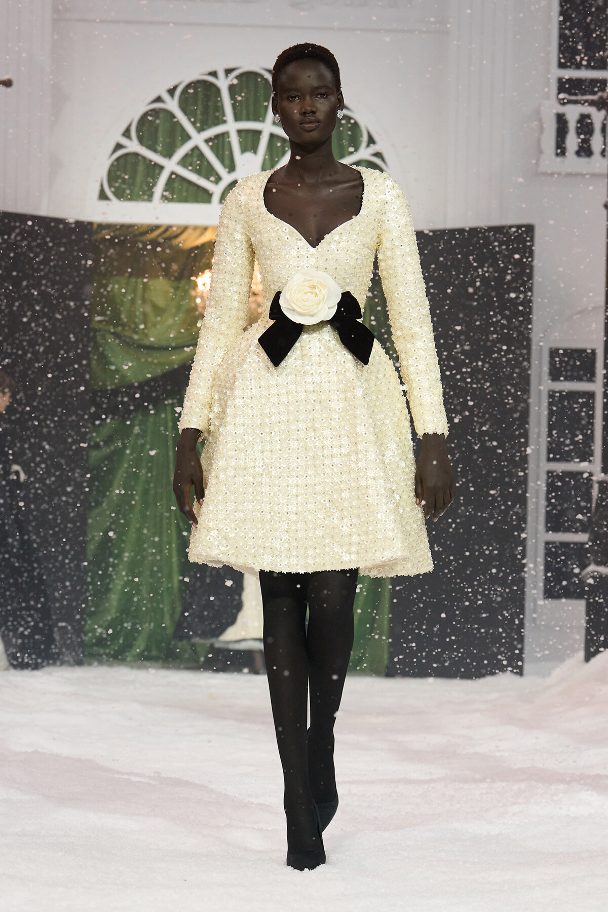 Model walking the runway in a cream beaded dress with a deep V neckline and a large white rose belt with black ribbons, snow-like backdrop and falling faux snow behind her.