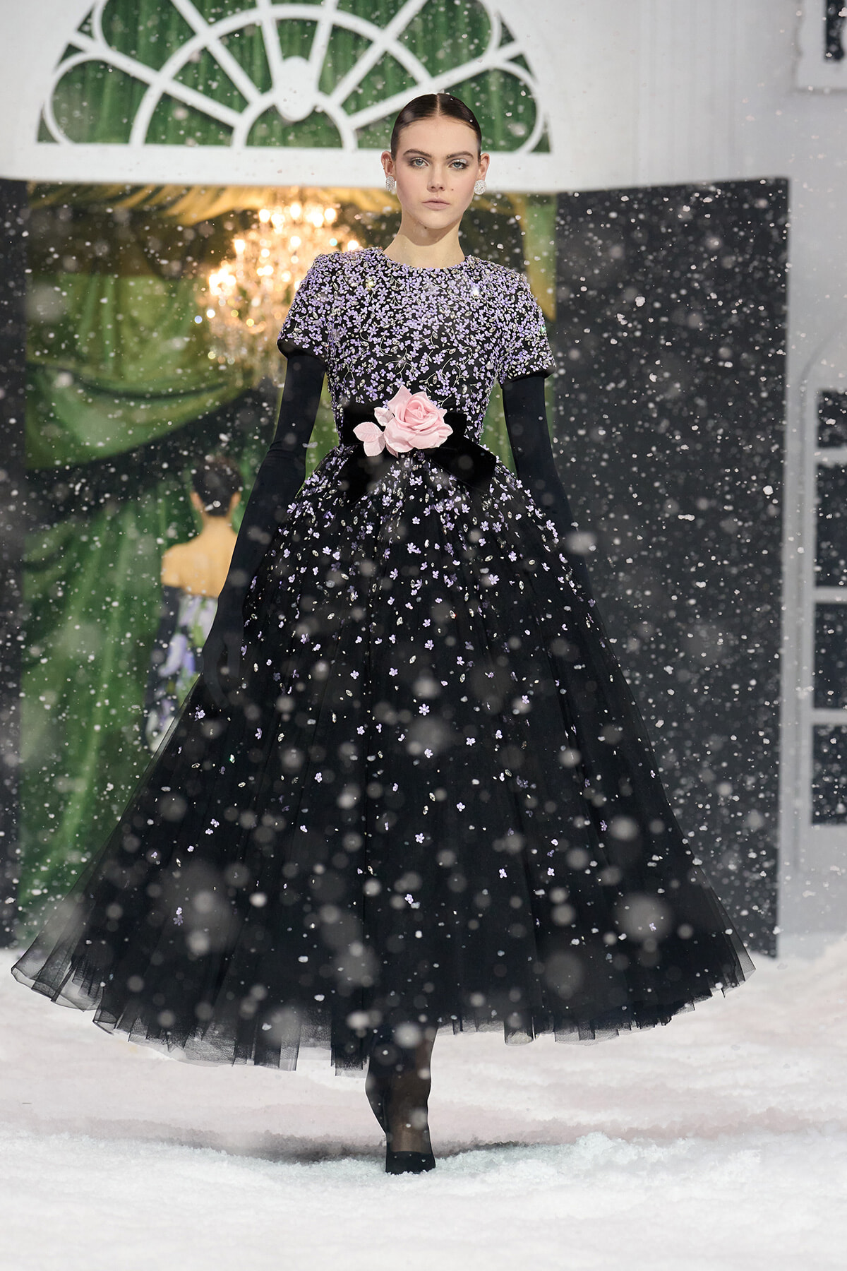 Model wearing a black, floor-length, sequin-embellished gown with pink rose belt, walking in a snowy fashion show.
