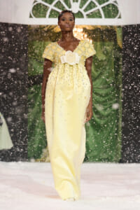 African American model in a pale yellow, bead‑embellished gown with a large ivory flower at the waist walking down a snow‑covered runway as snow falls.