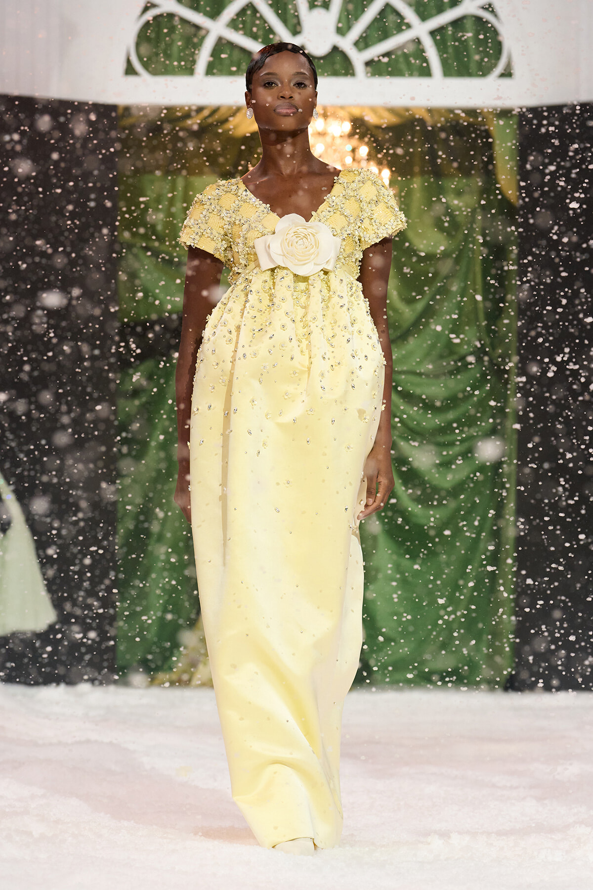 African American model in a pale yellow, bead‑embellished gown with a large ivory flower at the waist walking down a snow‑covered runway as snow falls.
