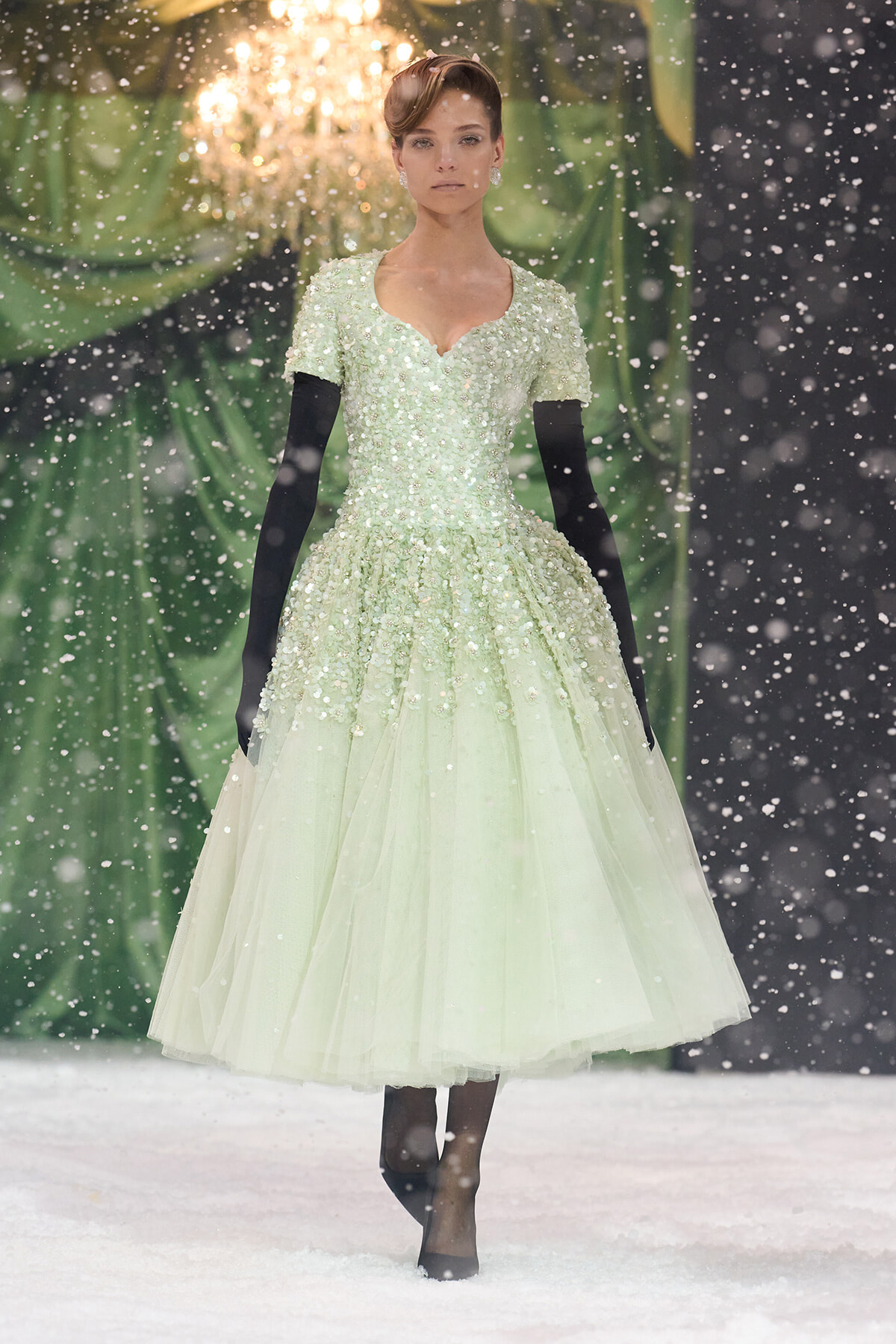 Model wearing a pale green, beaded ball gown with short sleeves and black opera gloves on a snowy runway backdrop.