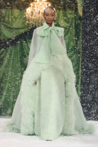 Model wearing a mint green gown with a large bow, sheer cape, and feathered trim walking in a snowy runway scene.