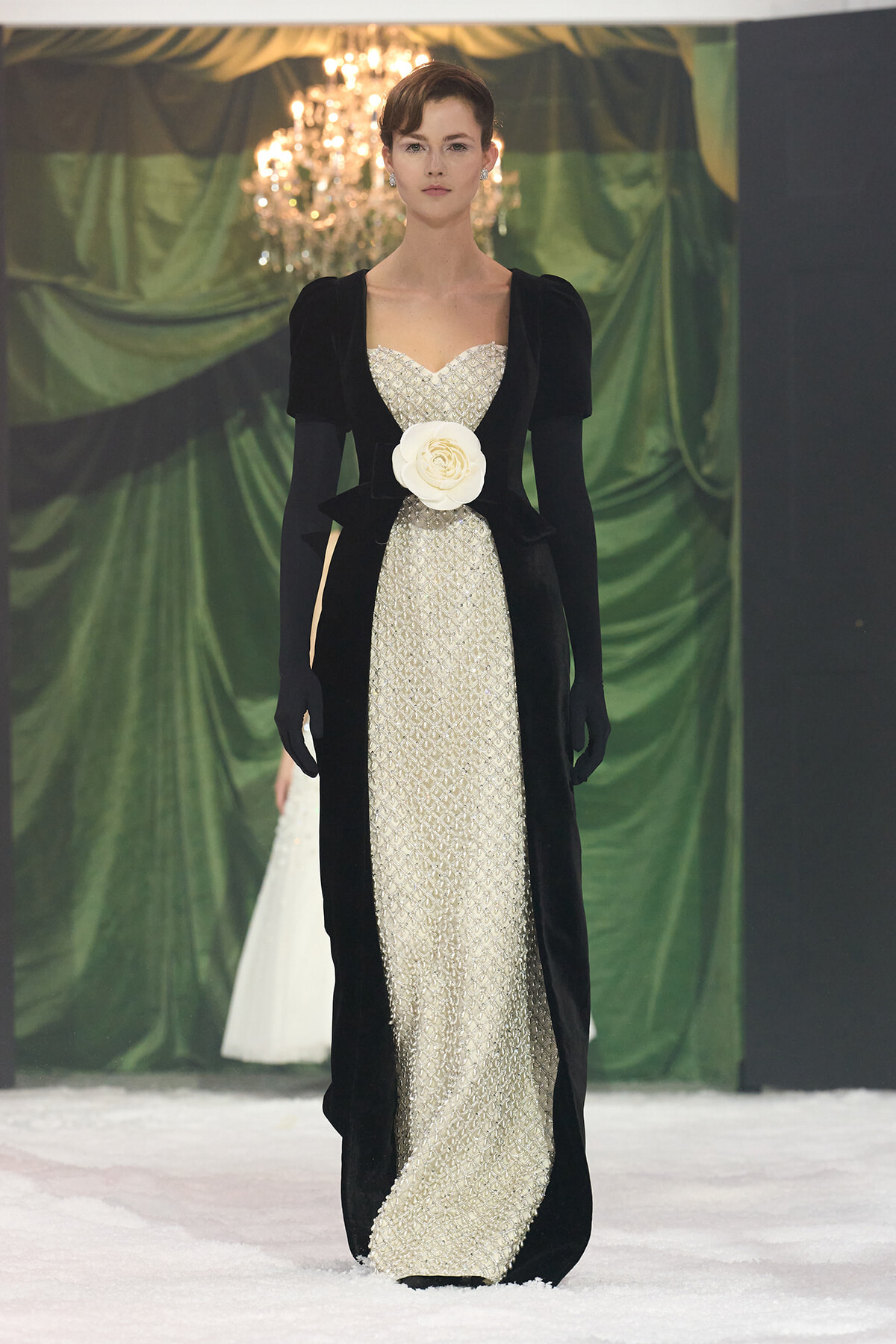 Model on a runway wearing a cream beaded gown with a black velvet overcoat and a large white flower belt, chandeliers in the background.
