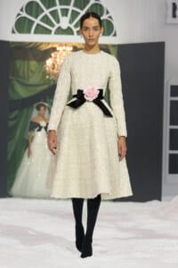 Model on runway wearing a cream textured midi dress with a pink rose belt and black ribbons, black tights, and pearl earrings.