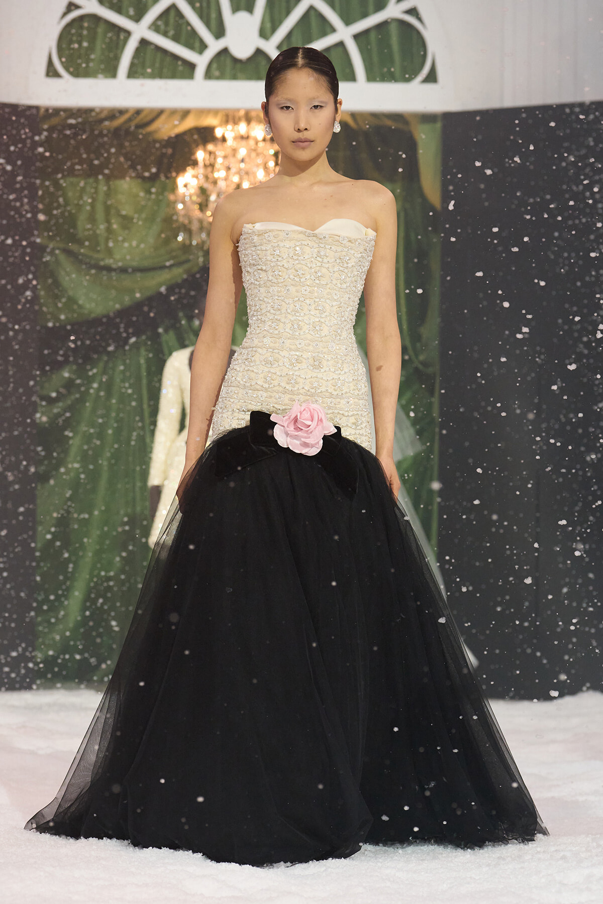 Model on a snowy runway wearing a strapless ivory beaded gown with a black tulle skirt and a pink flower at the waist.