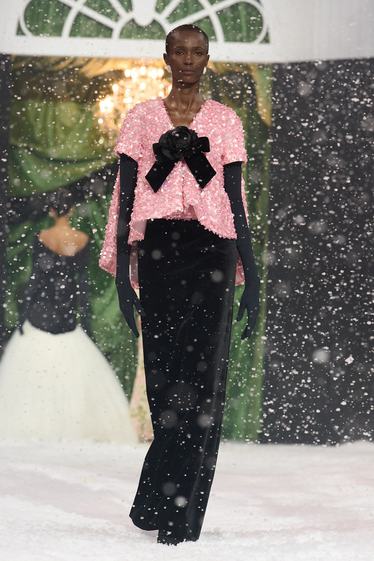 Model walking a snowy runway in a pink sequin top, black bow, and long black gloves.
