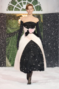 Model walking in a black and blush pink gown with a large pink rose belt, snowy backdrop, black gloves, and off-the-shoulder neckline