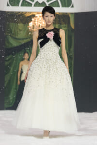Model walking a snowy runway in a white beaded ball gown with a black lace bodice and a pink rose at the chest.