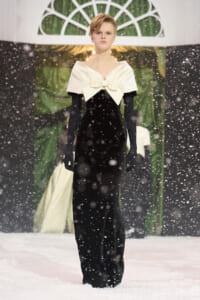 Model on a snowy runway wearing a black velvet gown with a large ivory bow at the neck and long black gloves.
