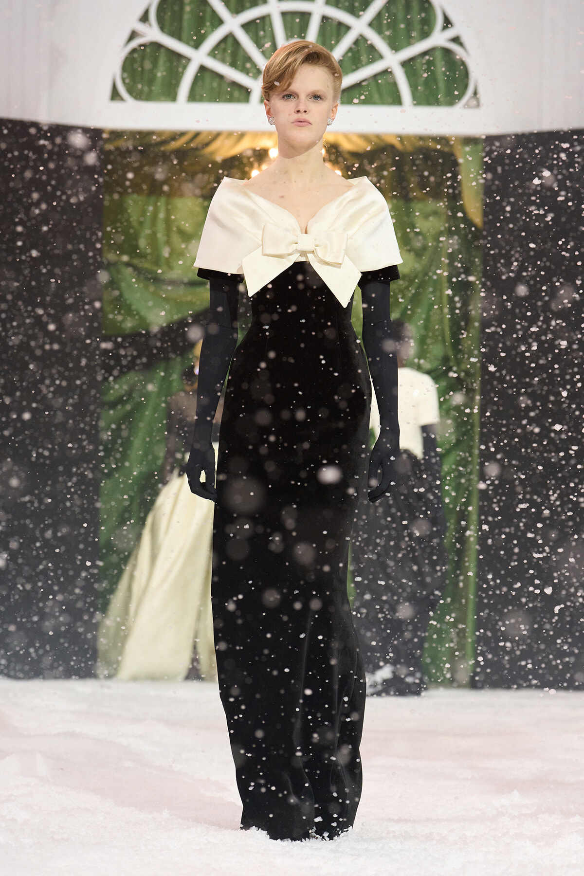 Model on a snowy runway wearing a black velvet gown with a large ivory bow at the neck and long black gloves.