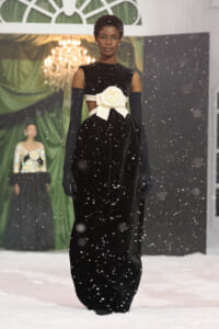 Model on a snowy runway wearing a long black velvet gown with off-the-shoulder sleeves, long gloves, and a large white bow at the waist