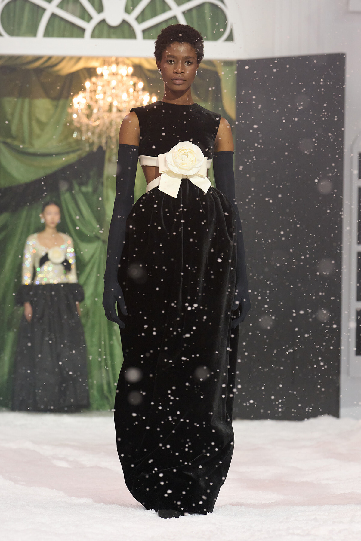 Model on a snowy runway wearing a long black velvet gown with off-the-shoulder sleeves, long gloves, and a large white bow at the waist